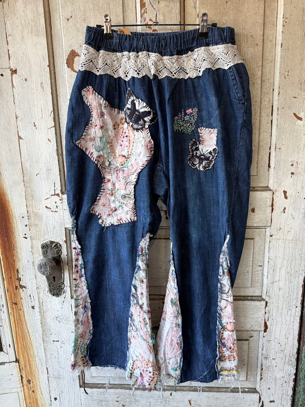 A pair of handmade, patchwork-style blue denim pants hanging on a rustic wooden door. The pants feature a cream lace waistband, floral patchwork inserts, and distressed, frayed hems.
