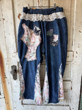 A pair of handmade, patchwork-style blue denim pants hanging on a rustic wooden door. The pants feature a cream lace waistband, floral patchwork inserts, and distressed, frayed hems.