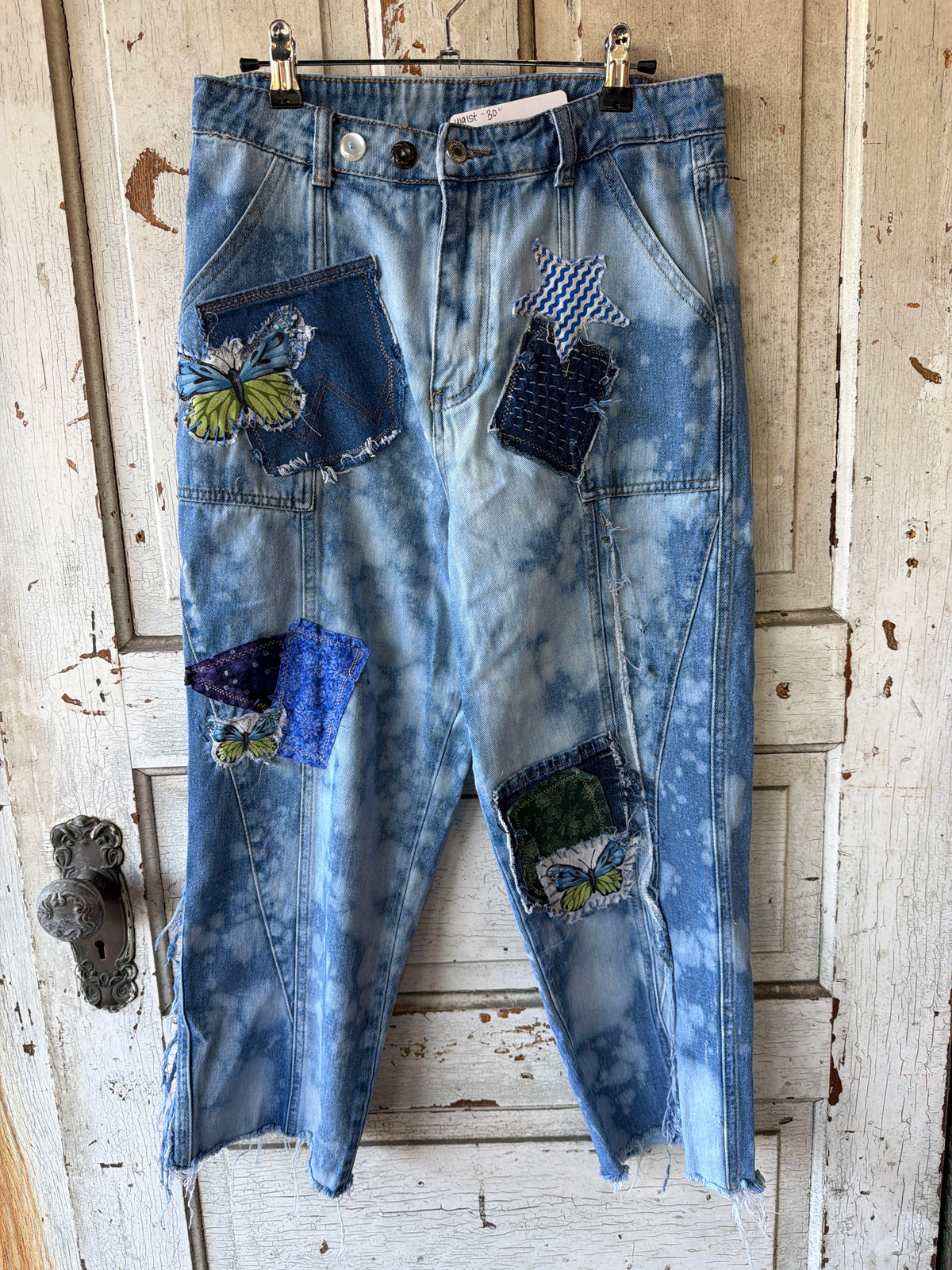 Tie-dye blue denim cropped pants with butterfly and patchwork applique details, hanging on a black hanger against a distressed white wooden door.