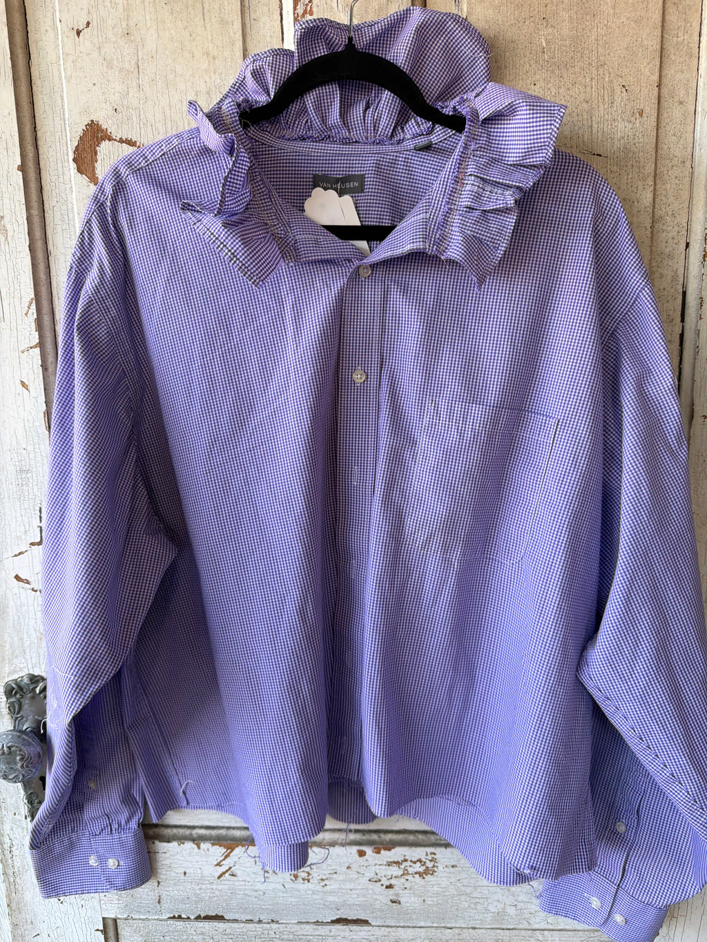 A lavender and white micro-checkered long-sleeve button-up shirt with a ruffled collar, hanging on a black hanger against a rustic, weathered wooden door.