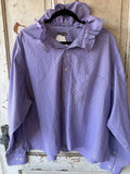 A lavender and white micro-checkered long-sleeve button-up shirt with a ruffled collar, hanging on a black hanger against a rustic, weathered wooden door.