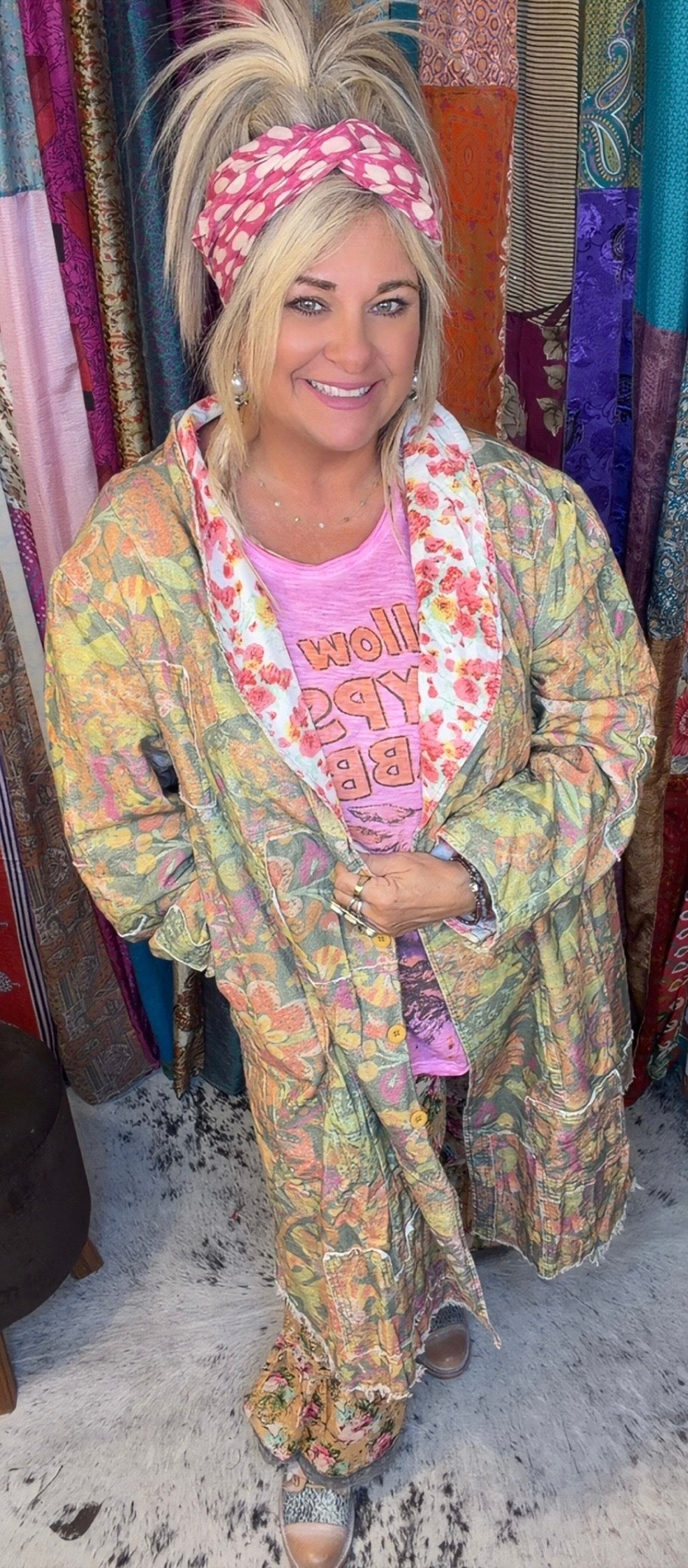 A woman wearing a colorful, patterned bohemian-style robe over a pink graphic t-shirt and a pink patterned headband, standing in front of a colorful fabric display.