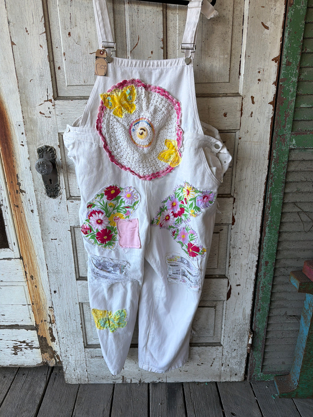 White denim bib overalls with colorful floral and crochet appliques, hanging on a rustic white wooden door.