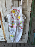 White denim bib overalls with colorful floral and crochet appliques, hanging on a rustic white wooden door.