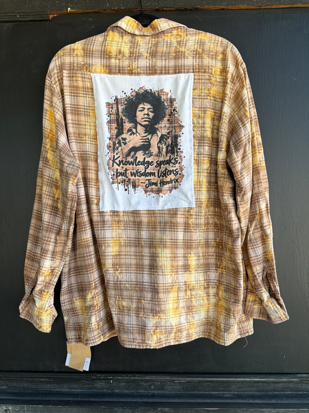 A long-sleeve plaid flannel shirt in brown, beige, and yellow tones, shown hanging against a dark background. The back of the shirt features a large white rectangular graphic patch with a portrait of Jimmie Ford and the quote 'Knowledge speaks, but wisdom listens.
