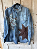 A light blue denim western-style shirt jacket with two large star patches, hanging on a black hanger against a distressed white wooden door. One star patch is brown and floral, and the other is solid dark brown.