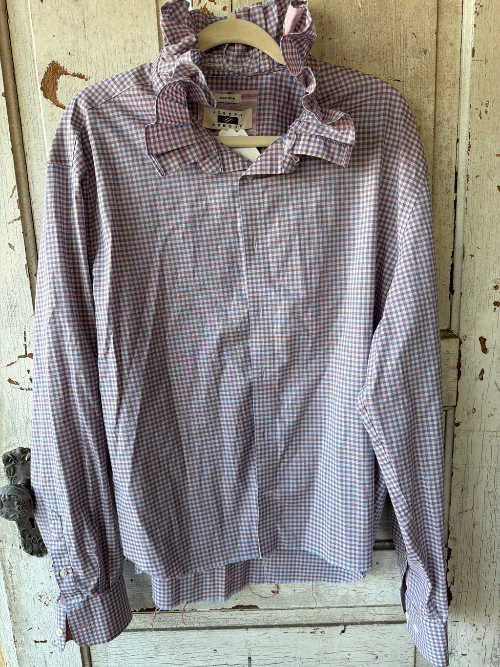 A long-sleeve button-up shirt with a pink and blue gingham check pattern and a ruffled collar, hanging on a wooden hanger against a distressed white wooden door.