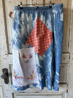 A handmade patchwork skirt in blue denim and white fabric, featuring a coral-red floral diamond patch, a white patch with colorful embroidered butterflies, and a blue elastic waistband, hanging on a rustic wooden door.