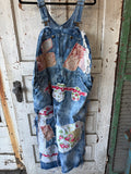 A pair of vintage-style blue denim patchwork overalls hanging on a black hanger against a distressed white wooden door. The overalls feature various colorful patchwork fabric inserts with floral and paisley patterns.