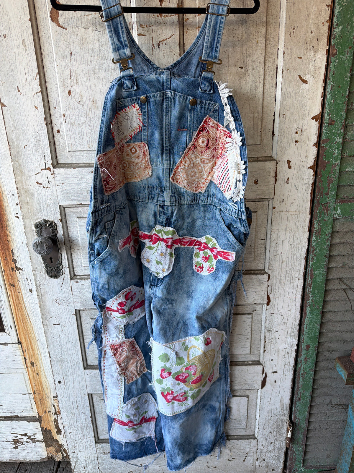 A pair of vintage-style blue denim patchwork overalls hanging on a black hanger against a distressed white wooden door. The overalls feature various colorful patchwork fabric inserts with floral and paisley patterns.