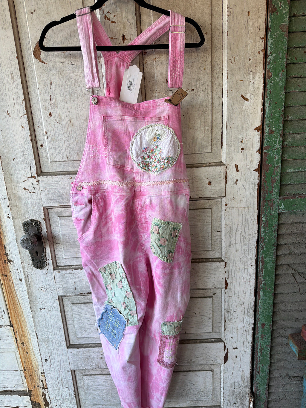 Pink patchwork patchwork overall hanging on a black hanger against a distressed white wooden door. The overall features pink watercolor wash fabric with various patchwork pockets in green, blue, and floral prints.