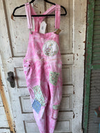 Pink patchwork patchwork overall hanging on a black hanger against a distressed white wooden door. The overall features pink watercolor wash fabric with various patchwork pockets in green, blue, and floral prints.