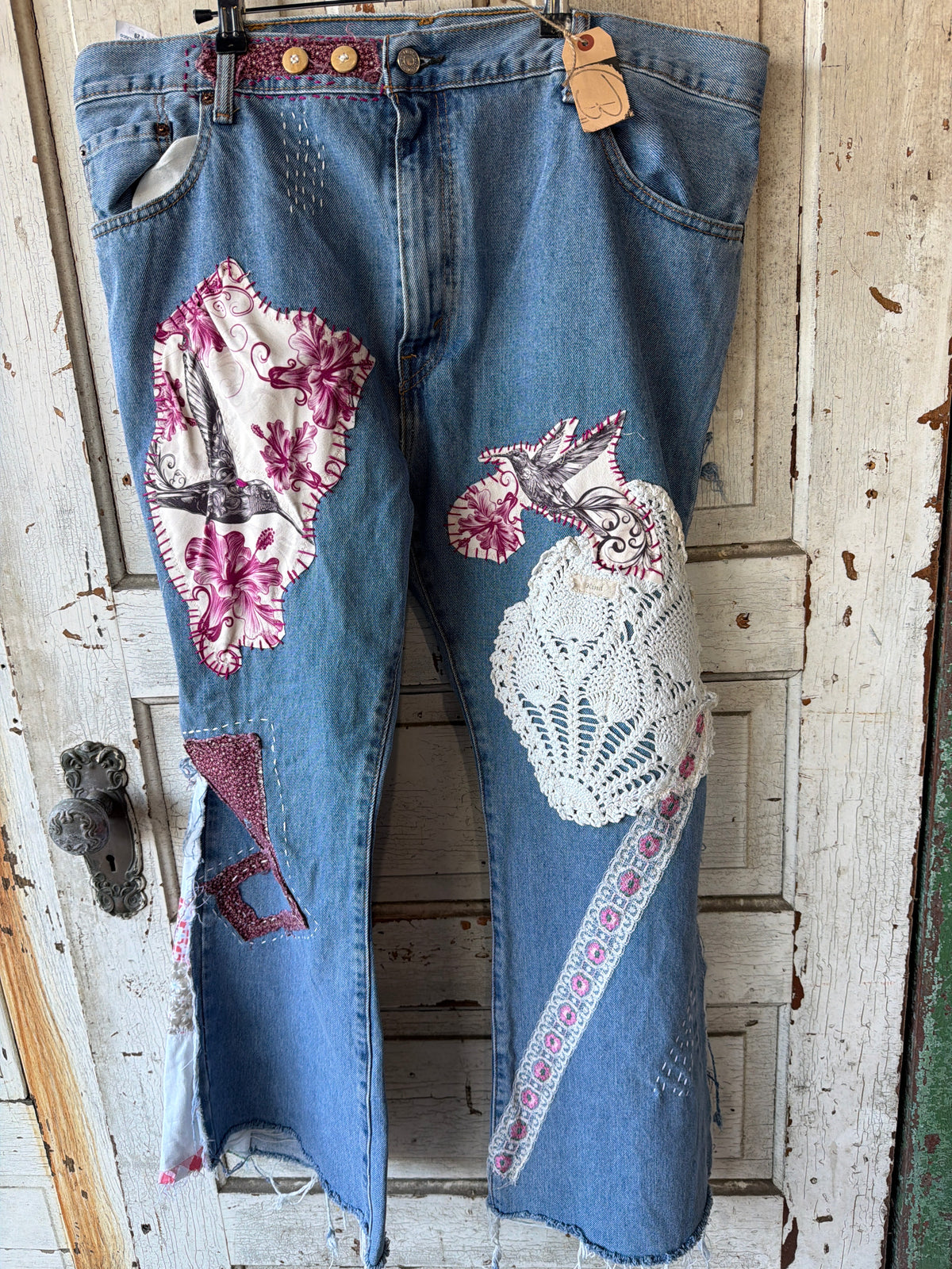 Distressed blue denim jeans with bohemian patchwork details, including floral fabric patches, crochet applique, and embroidery, hanging on a rustic white wooden door.
