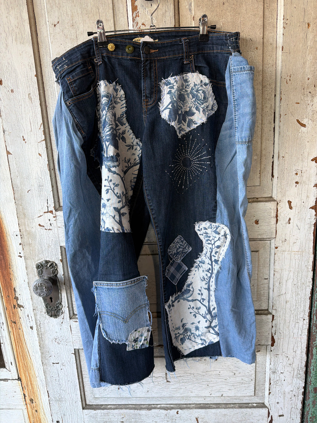 Patchwork denim capri pants hanging on a metal hanger against a distressed white wooden door. The dark blue denim features light blue denim panels, floral blue-and-white patterned fabric patches, and a blue sunburst applique.