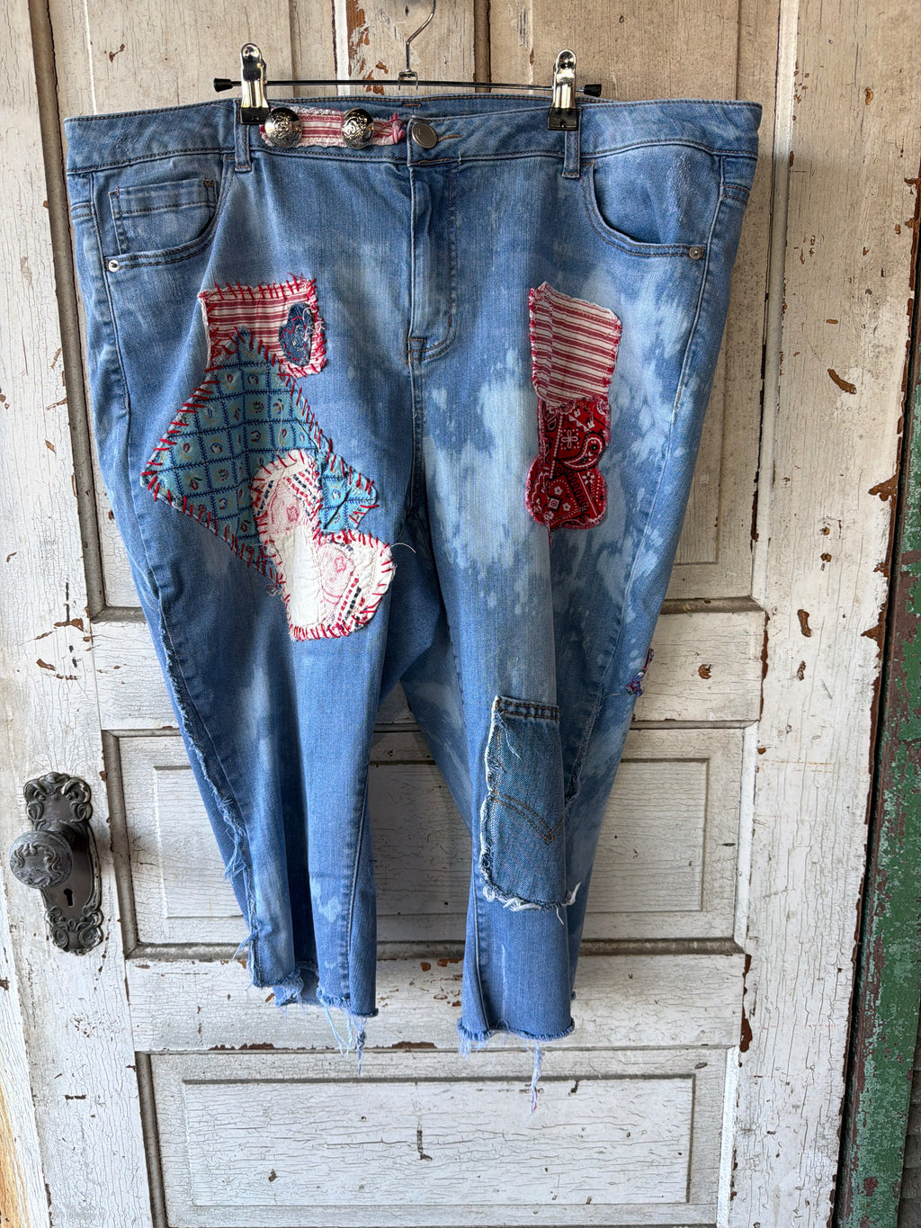 Light blue denim jeans with a tie-dye wash and patriotic patchwork details, hanging on a metal hanger against a distressed white wooden door.