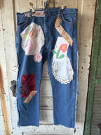 A pair of blue denim jeans with extensive hand-painted and embroidered patchwork details, hanging on a rustic wooden door.