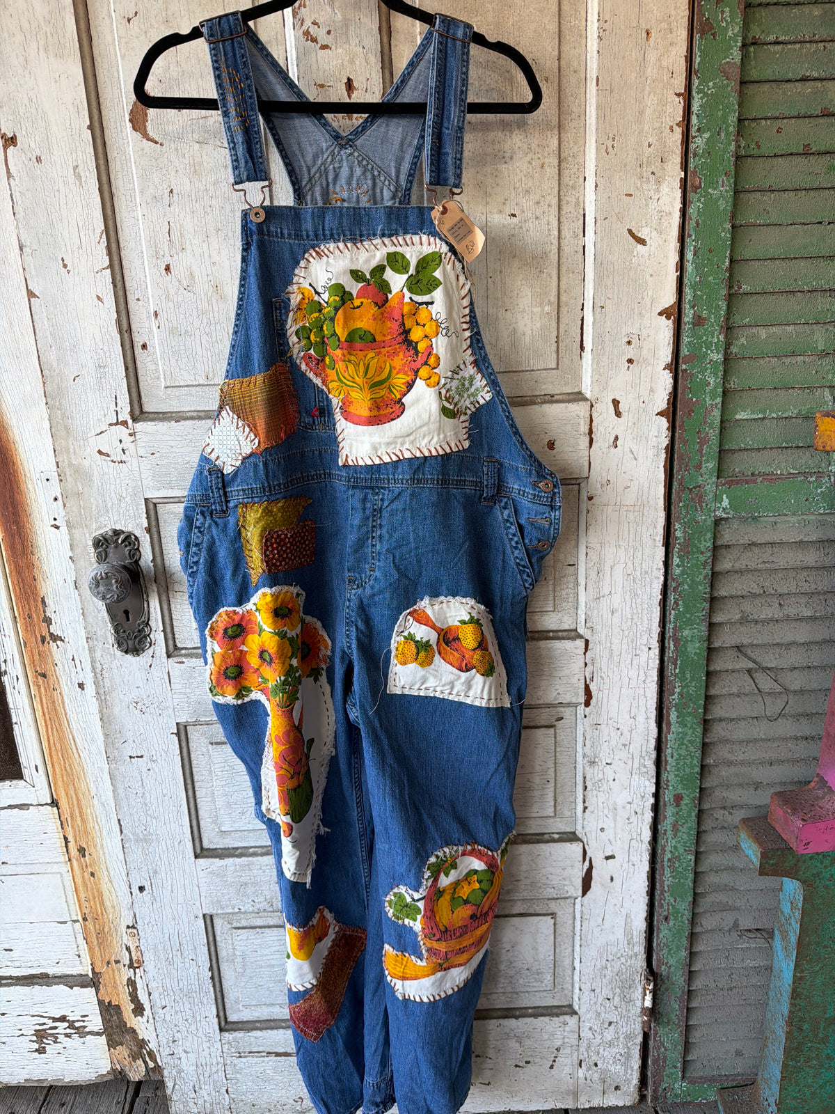 A pair of blue denim bib overalls hanging on a black hanger against a distressed white wooden door. The overalls feature colorful embroidered patches depicting autumn harvest motifs, including pumpkins, gourds, and flowers, on the bib and legs.