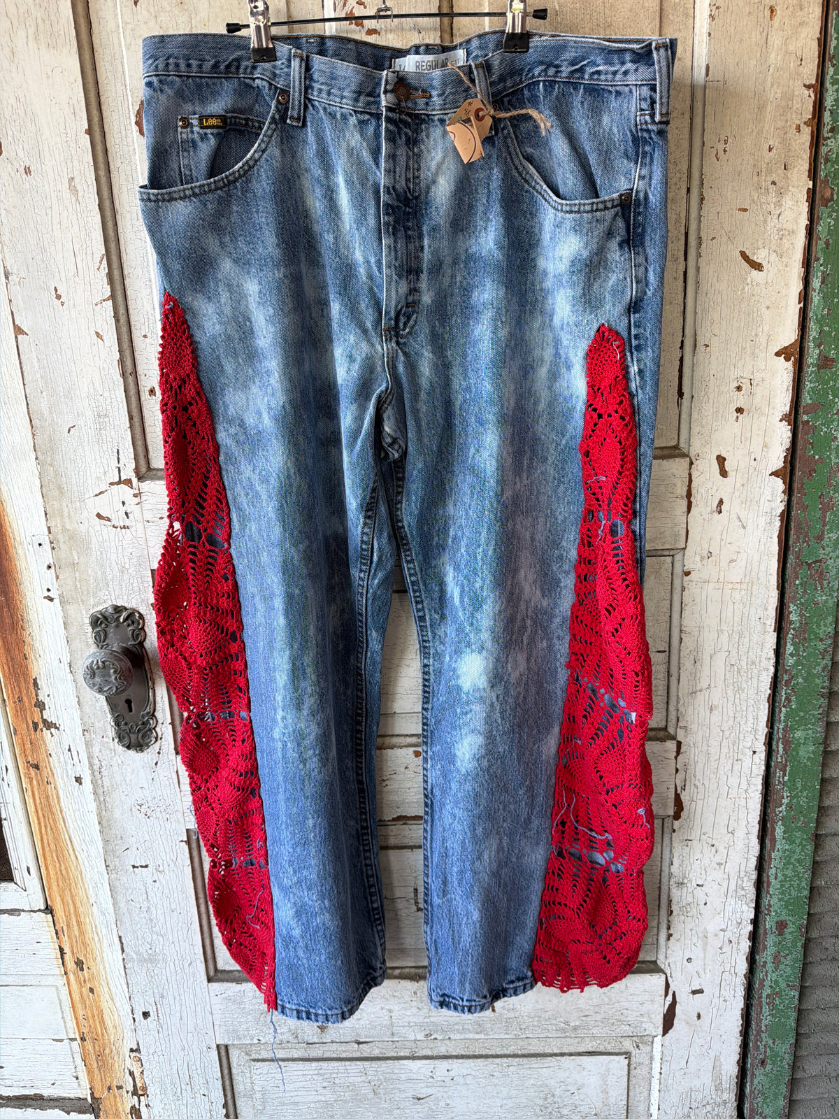 Light blue acid-wash denim jeans with bright red crochet lace panels on the lower legs, hanging on a metal hanger against a distressed white wooden door.