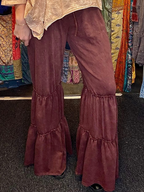 A person wearing wide-leg flared pants in a dusty rose or mauve-brown velvet-like fabric, paired with a light patterned top. The photo is cropped from the waist down, showing the pants hanging or worn in what appears to be a boutique setting with colorful garments in the background.