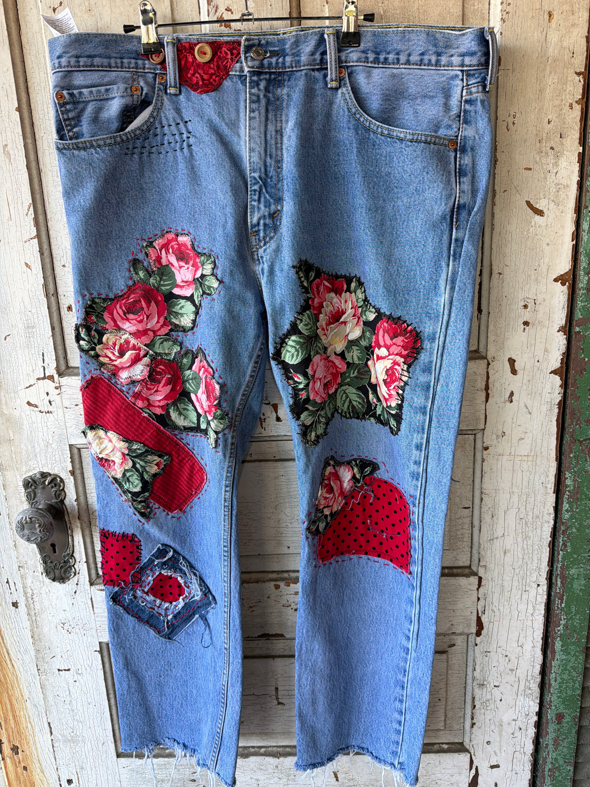Light blue denim jeans with large embroidered rose patches and red fabric accents, hanging on a hanger against a distressed white wooden door.