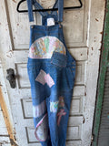 A pair of blue denim patchwork overalls hanging on a black hanger against a distressed white wooden door. The overalls feature various colorful patchwork fabric inserts on the front, including a pink cross, a pink bow, and a floral semicircle patch.