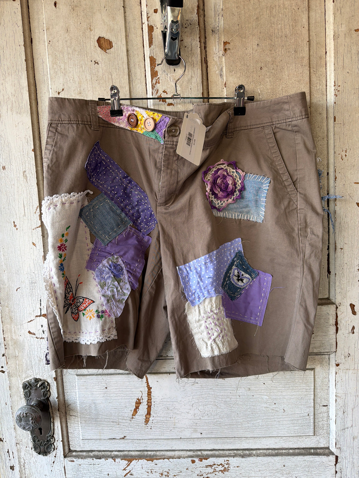 A pair of brown patchwork shorts hanging on a metal hanger against a distressed white wooden door. The shorts feature various colorful fabric patches sewn onto the front, including purple, blue, white, and floral designs.