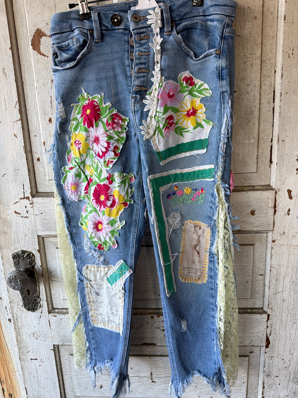 Light blue denim jeans with heavy distressing, frayed hems, and colorful floral patchwork appliques, hanging on a black hanger against a distressed white wooden door.