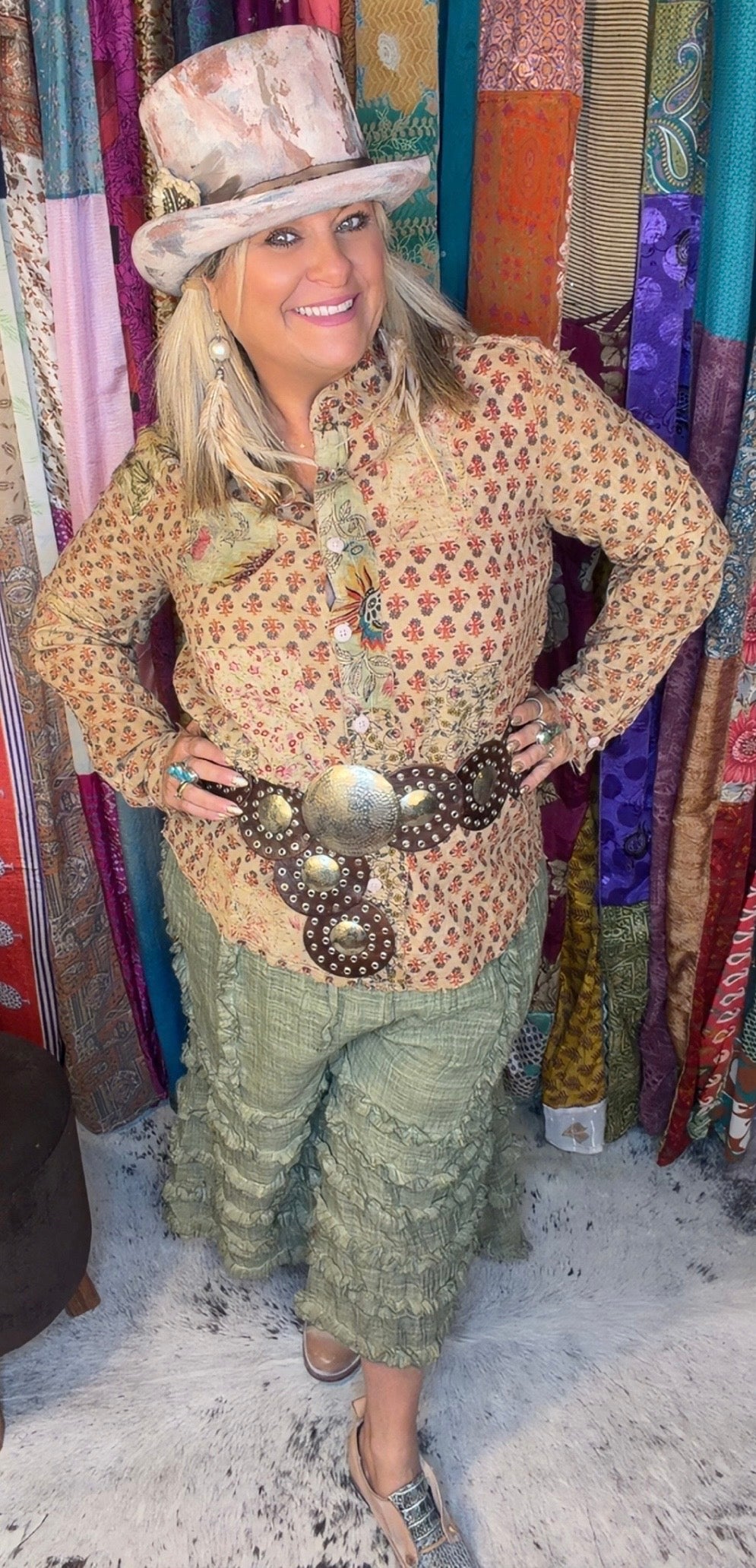 A woman posing in a bohemian-style outfit featuring a beige patterned long-sleeve top, sage green ruffled pants, a large silver concho belt, and a matching hat, set against a colorful background of hanging textiles.