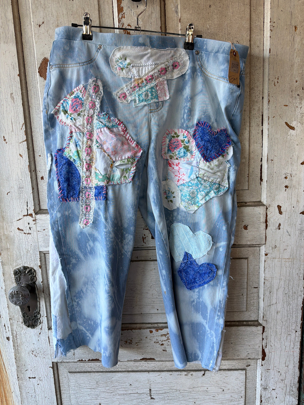 Light blue tie-dye cropped jeans with colorful patchwork appliques, including hearts and floral fabrics, hanging on a rustic white wooden door.