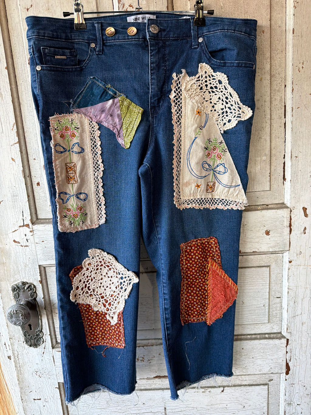 A pair of blue denim jeans with extensive handmade patchwork embellishments, including lace doilies, embroidered fabric panels, and patterned fabric patches, hanging on a rustic white wooden door.