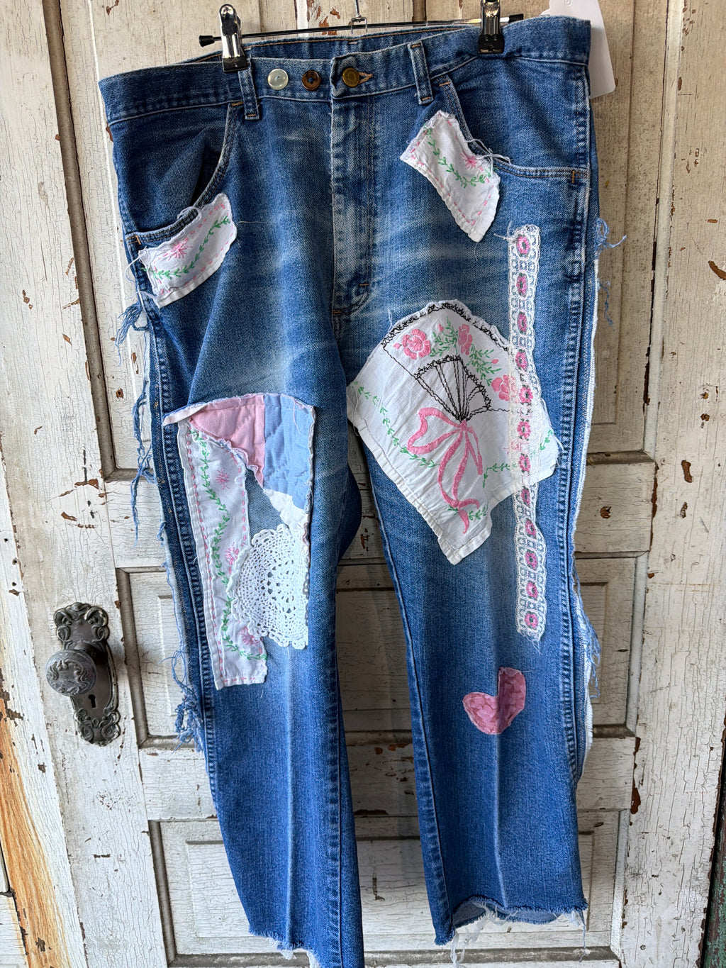 A pair of blue denim jeans with distressed frayed hems and multiple colorful patchwork fabric appliques, hanging against a rustic white wooden door.