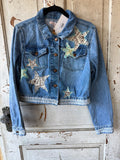 A cropped blue denim jacket with star patches, hanging on a black hanger against a distressed white wooden door. The jacket features brown buttons, a button-down front, and decorative star patches on the chest and pocket areas.