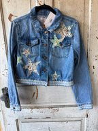 A cropped blue denim jacket with star patches, hanging on a black hanger against a distressed white wooden door. The jacket features brown buttons, a button-down front, and decorative star patches on the chest and pocket areas.