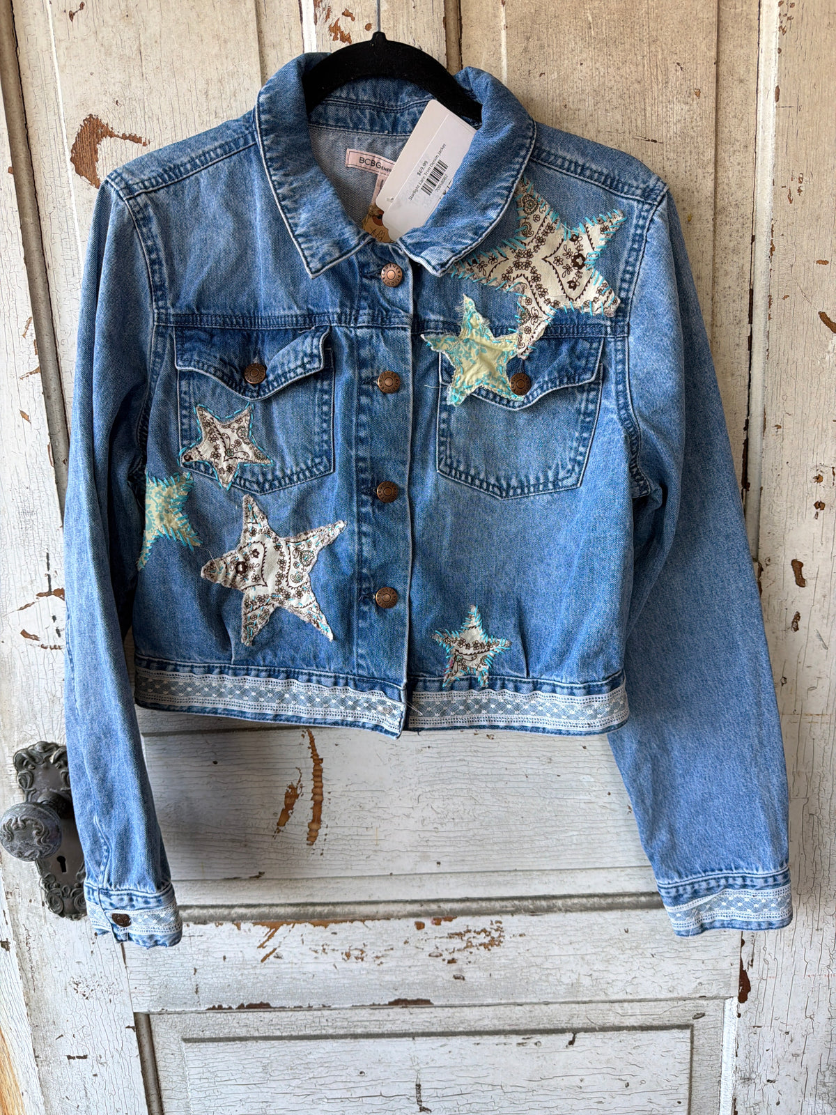 A cropped blue denim jacket with star patches, hanging on a black hanger against a distressed white wooden door. The jacket features brown buttons, a button-down front, and decorative star patches on the chest and pocket areas.
