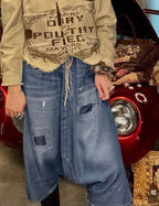 A woman stands in a cluttered boutique or garage-like shop setting wearing a beige distressed graphic shirt, a blue denim skirt, and a black beret. A red vintage car is behind her, and bags and clothing racks are visible around her.