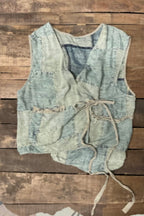 A sleeveless patchwork vest made from light blue denim and beige corduroy-like fabric, featuring a tie closure at the front, laid flat on a rustic wooden plank background.